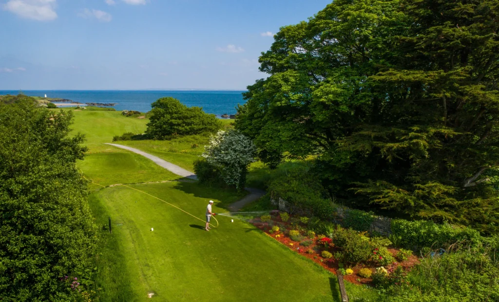 Groundskeeper watering flowerbeds by the tee on a coastal golf course with sea views and a distant lighthouse