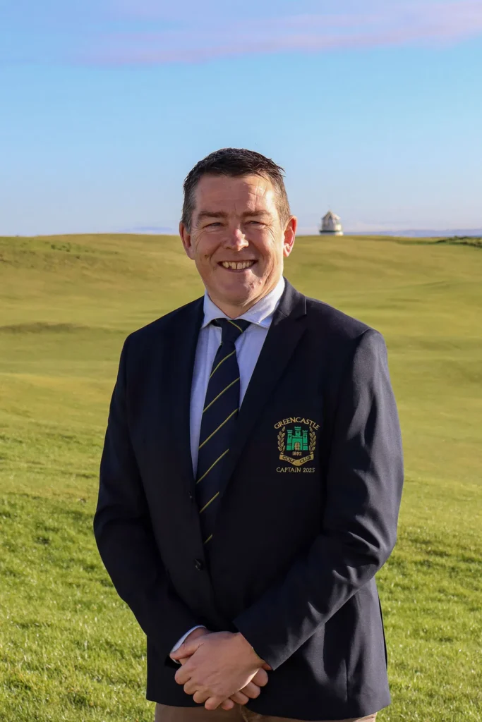 Smiling man in a navy blazer with Greencastle Golf Club crest on a sunny links fairway, distant dome behind