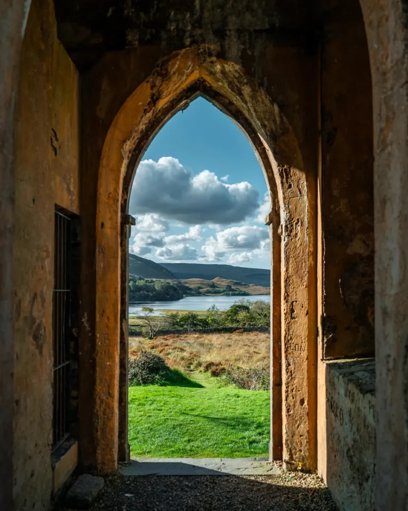 Gothic stone arch in a ruin framing an Irish lake, green fields and hills under bright clouds