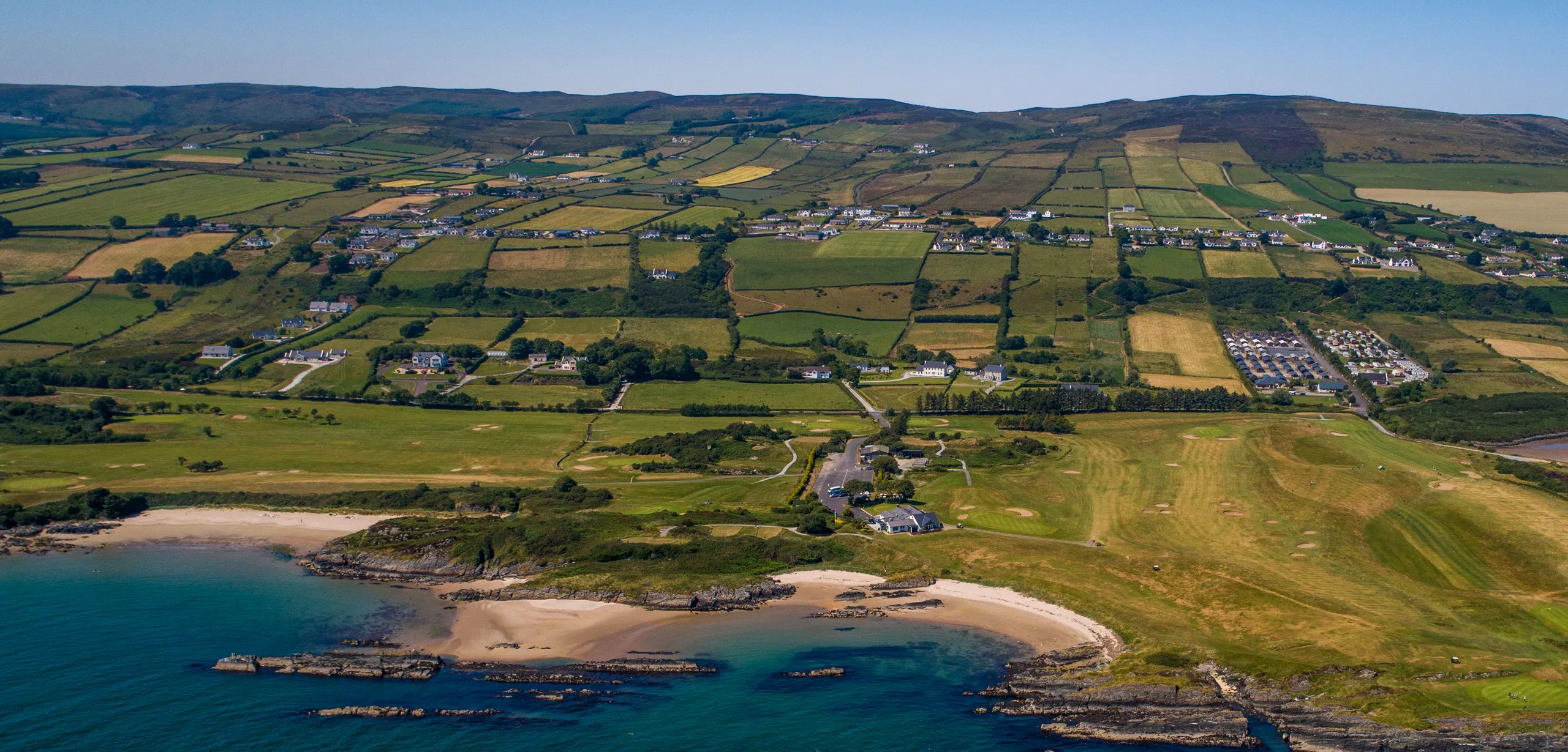Patchwork fields and a seaside golf course above sandy coves and blue water on the Irish coast