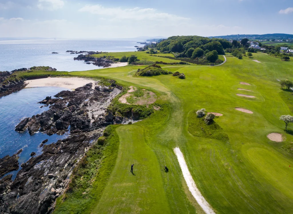 Aerial view of a coastal links course with rocky shore, sandy cove and a lone golfer on the fairway by the Atlantic.