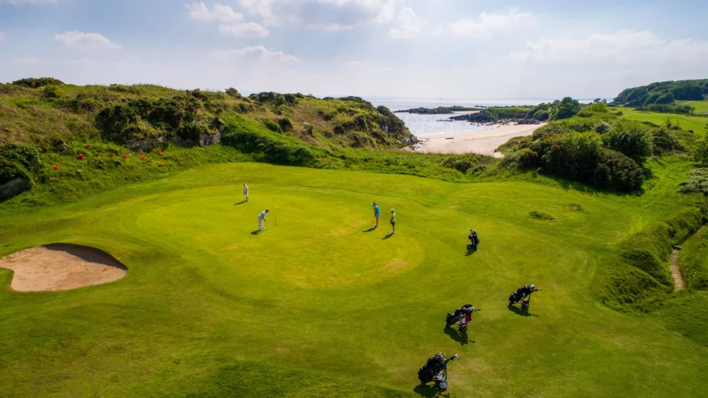 Golfers putting on a seaside links green, with a bunker and a sandy beach in the background under bright skies.