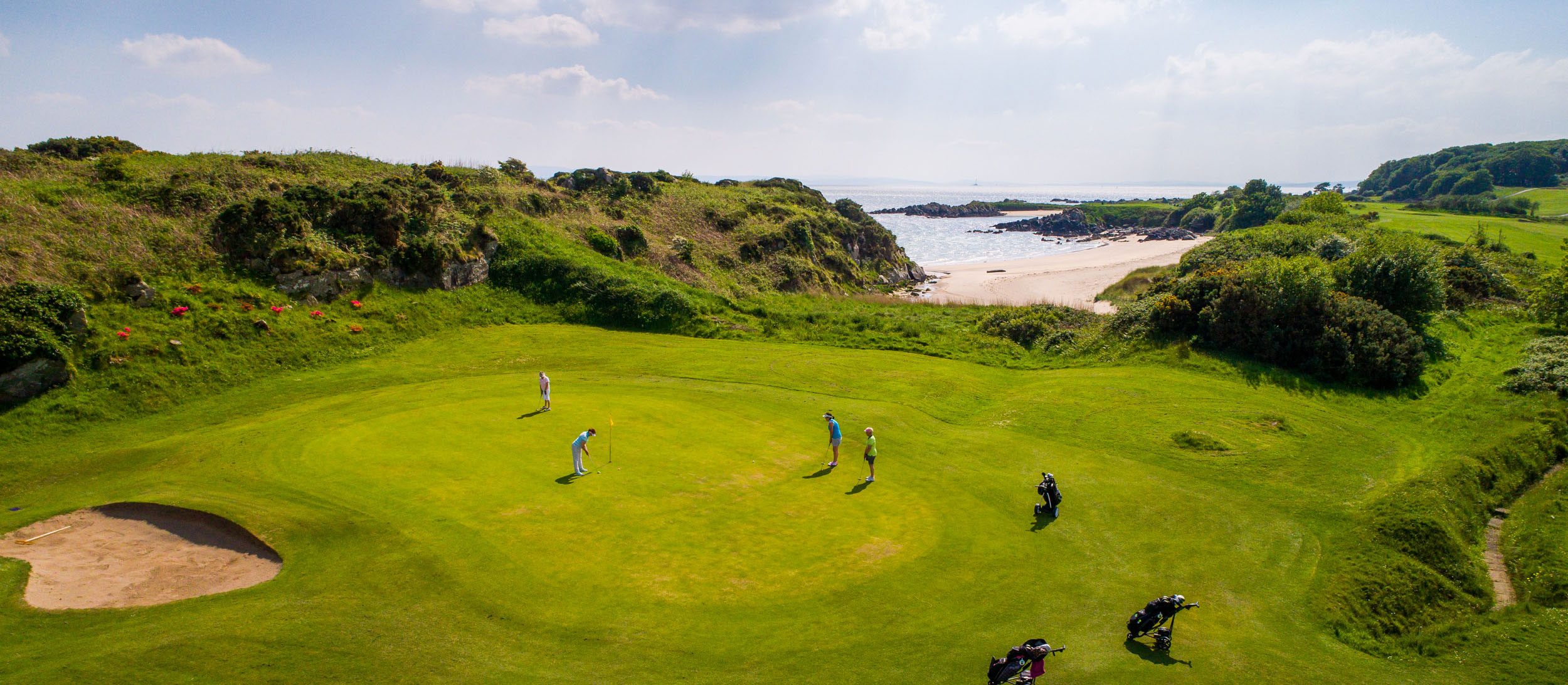 Golfers putting on a coastal links green beside a sandy cove on a bright day in Ireland