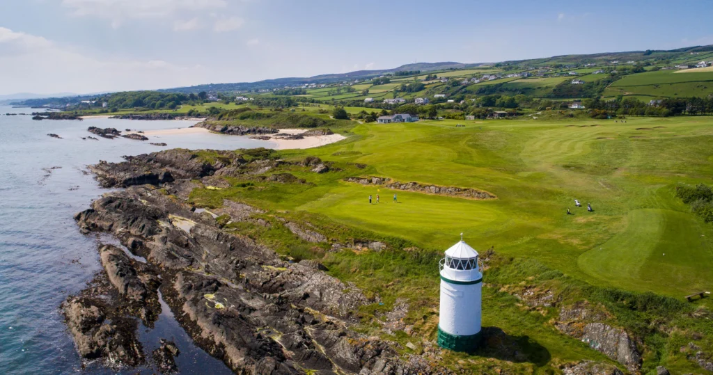 Clifftop lighthouse by a coastal links golf course, golfers on a fairway above rocky shore and a sandy cove.