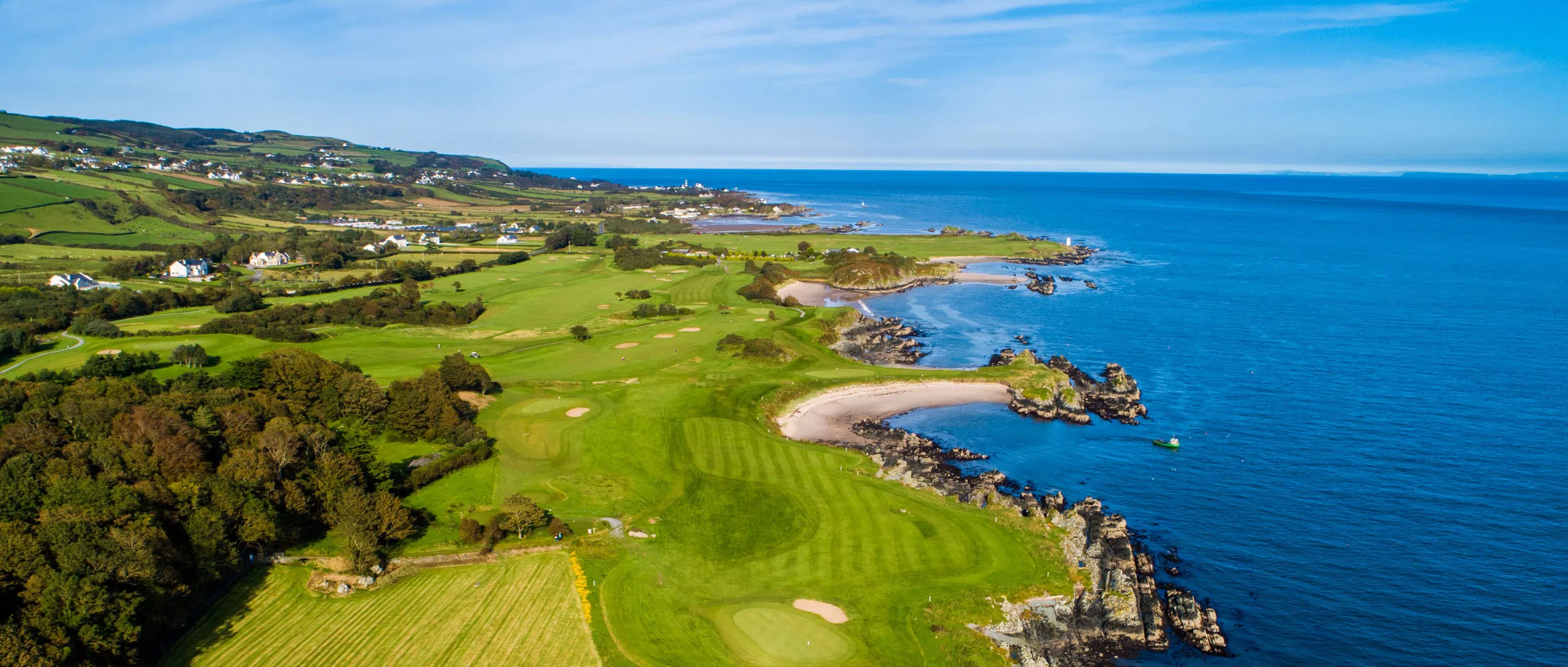 Aerial of a links golf course on the Irish coast, with rocky coves, bright fairways and a calm blue sea.