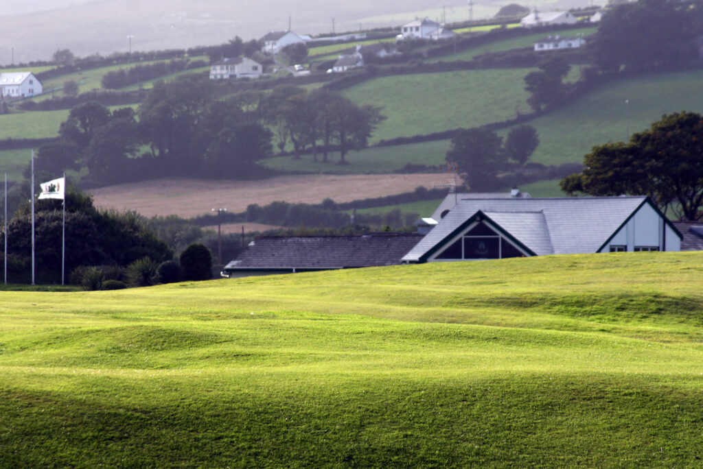 Rolling golf course fairway by a clubhouse and flagpoles, with patchwork fields and cottages beyond.