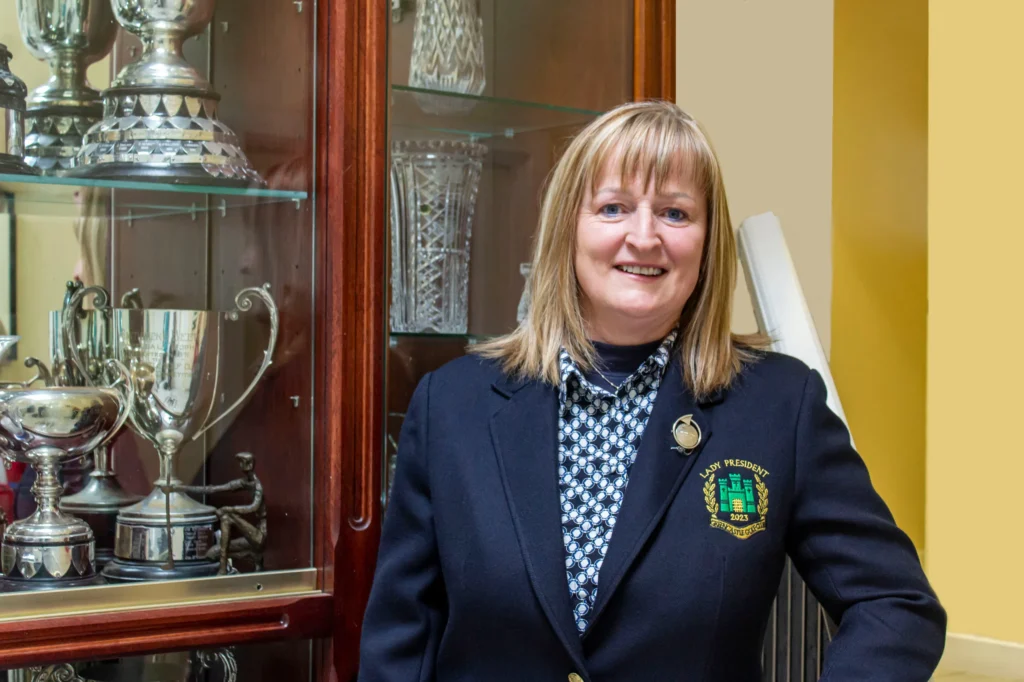 Smiling woman in a navy blazer with club crest beside a trophy cabinet of silver cups and crystal.