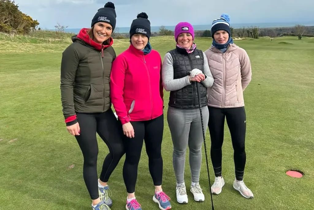Four women golfers in woolly hats smiling on a chilly links course with sea views in Ireland