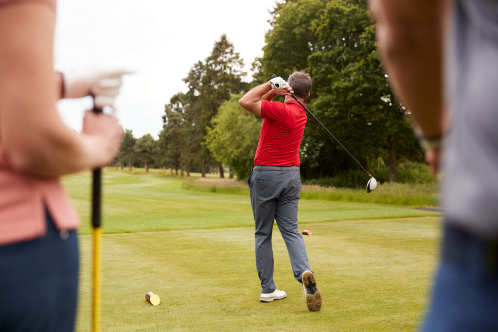 Golfer in a red shirt tees off on a parkland golf course while playing partners watch.