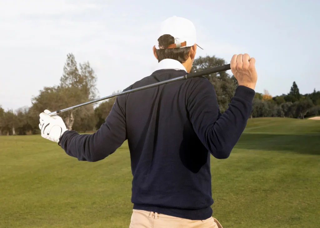 Golfer in navy jumper rests club across shoulders on a sunny golf course, white cap and glove.