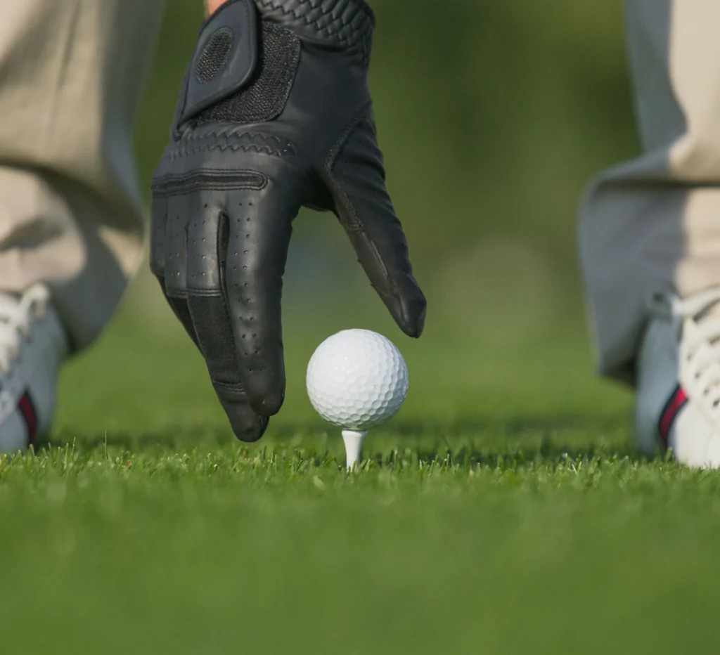 Golfer’s gloved hand tees up a golf ball on lush grass, ready for the tee shot.