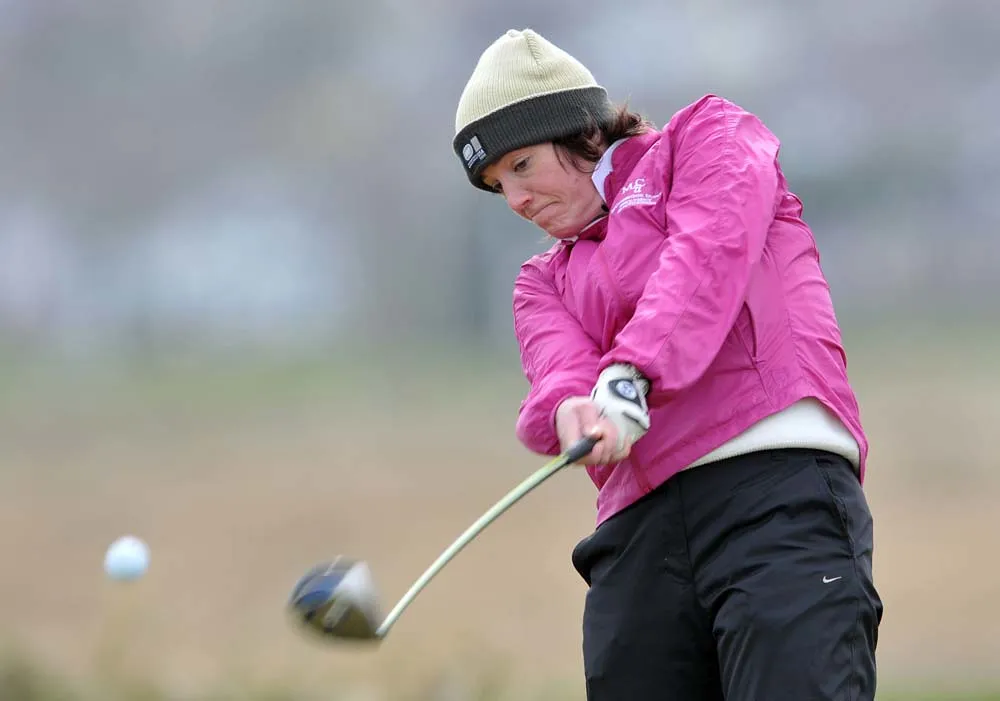 Golfer in a pink jacket and woolly hat drives a tee shot on a windy links course.