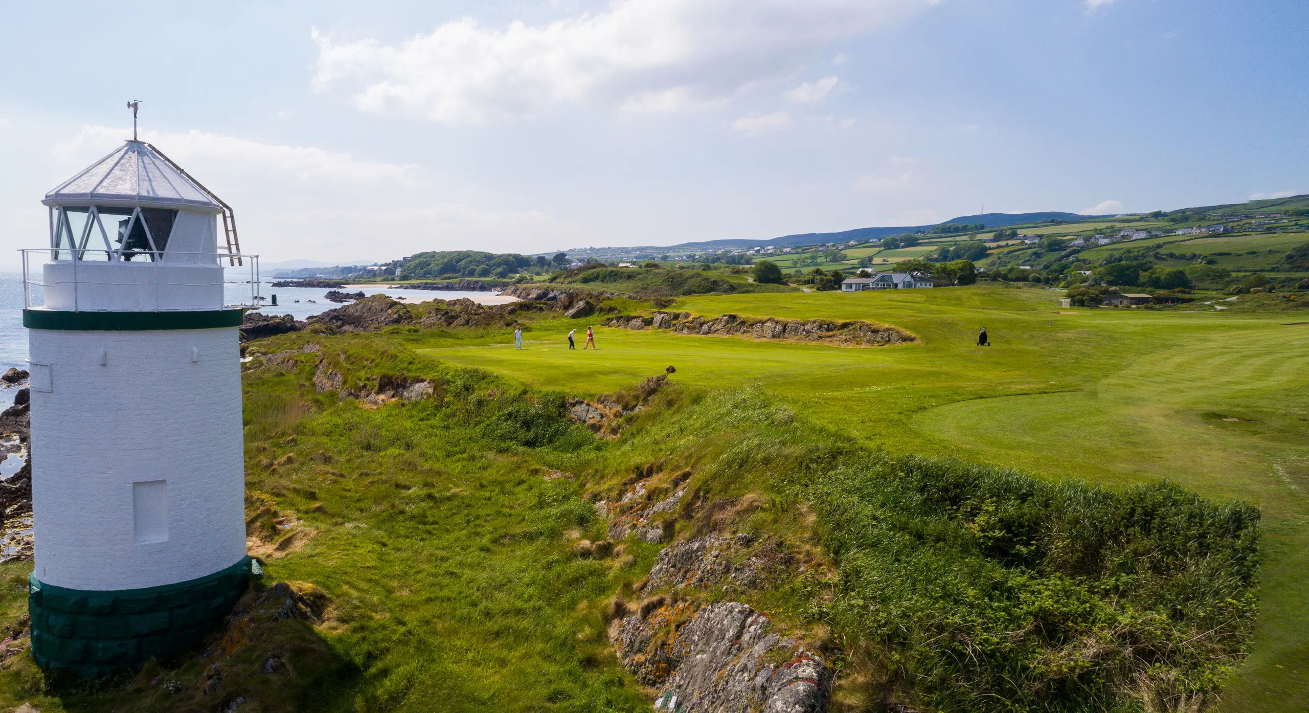 Coastal lighthouse beside a links golf course, golfers on the fairway with rolling Irish hills and sea beyond.