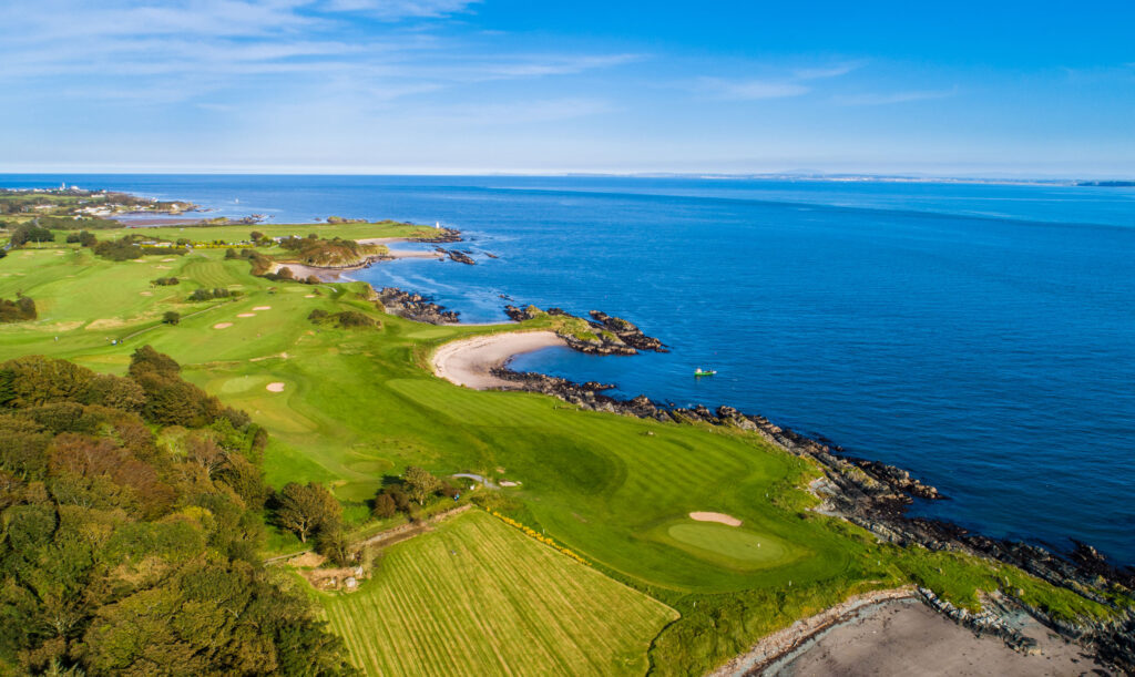 Coastal links golf course with rocky headlands, sandy coves and calm blue sea under a bright sky