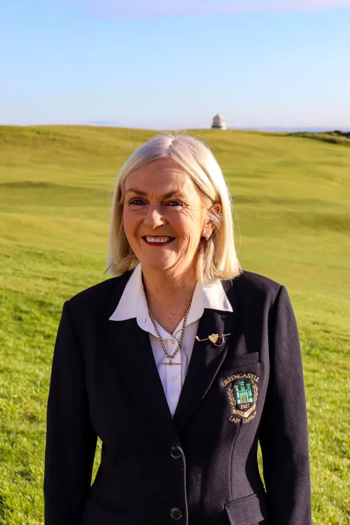 Smiling woman in Greencastle Golf Club blazer on a sunlit links, green fairways and blue sky behind