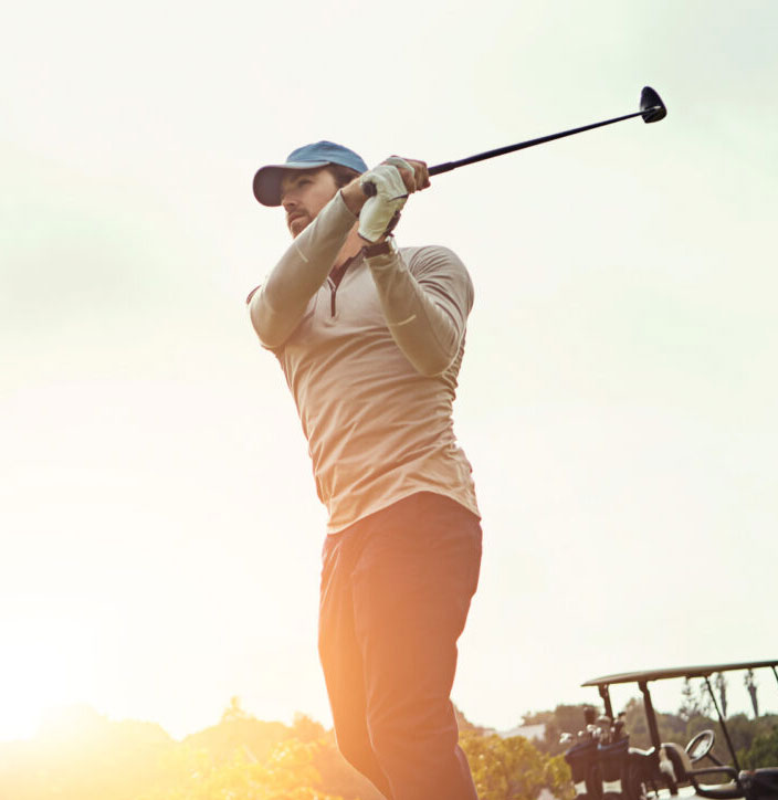 Golfer in cap finishing a smooth swing in warm evening light on a golf course, buggy in the background