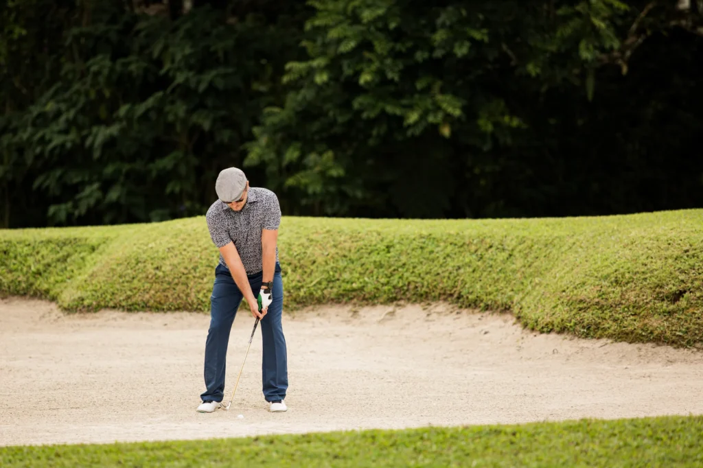 Golfer in a flat cap plays a bunker shot from the sand on a lush golf course