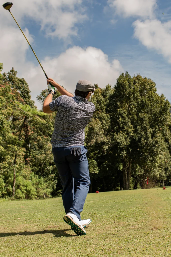Golfer follows through on a tee shot on a sunny golf course, ball soaring into blue sky.