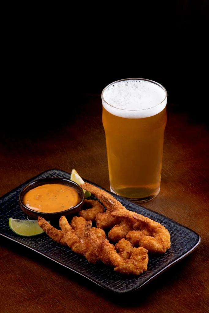 Crispy breaded prawns with spicy dipping sauce and lime wedges beside a frothy pint of lager.