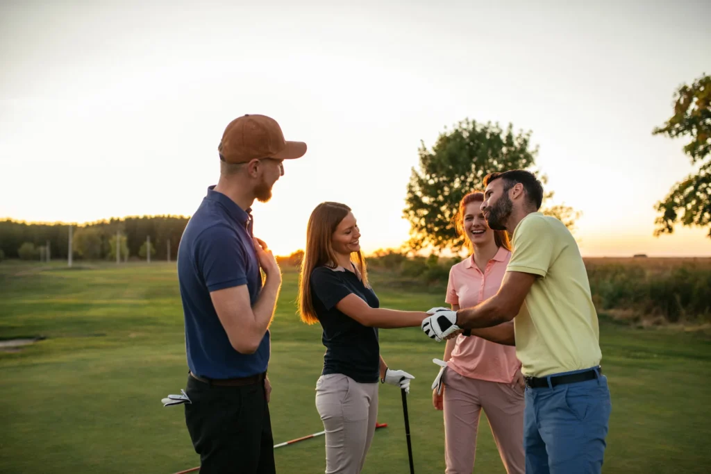 Four golfers share a handshake at sunset on the golf course, laughing with clubs after the round.