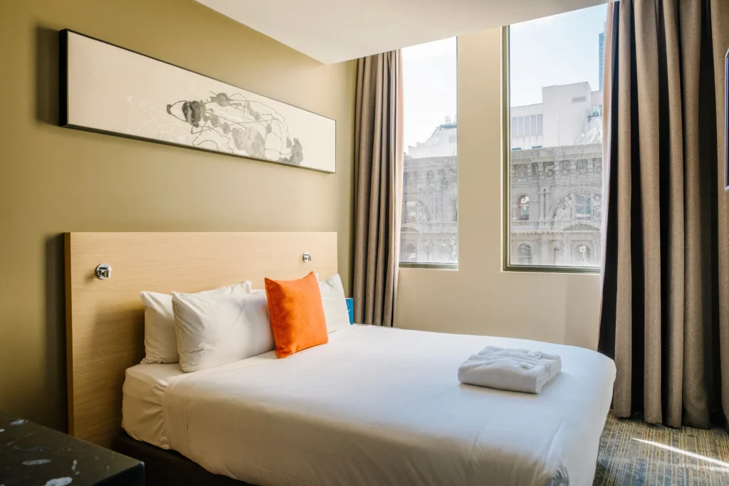 Bright hotel room with double bed, orange cushion and folded towel, large windows with a city view.