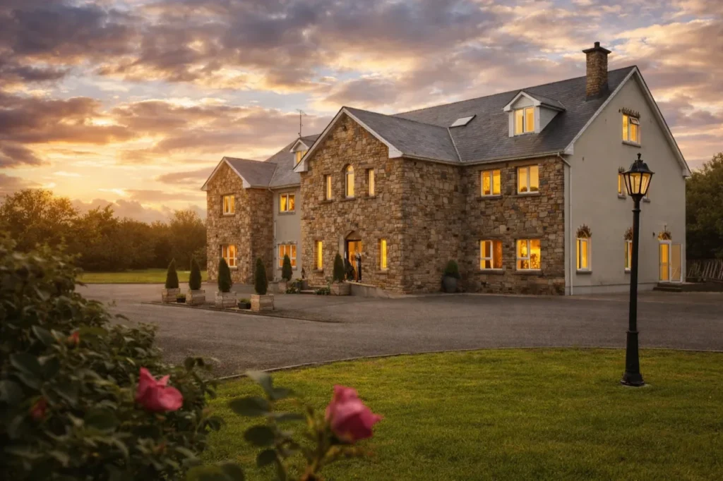 Warm-lit stone country house in rural Ireland at sunset, with roses and a lamppost by the drive