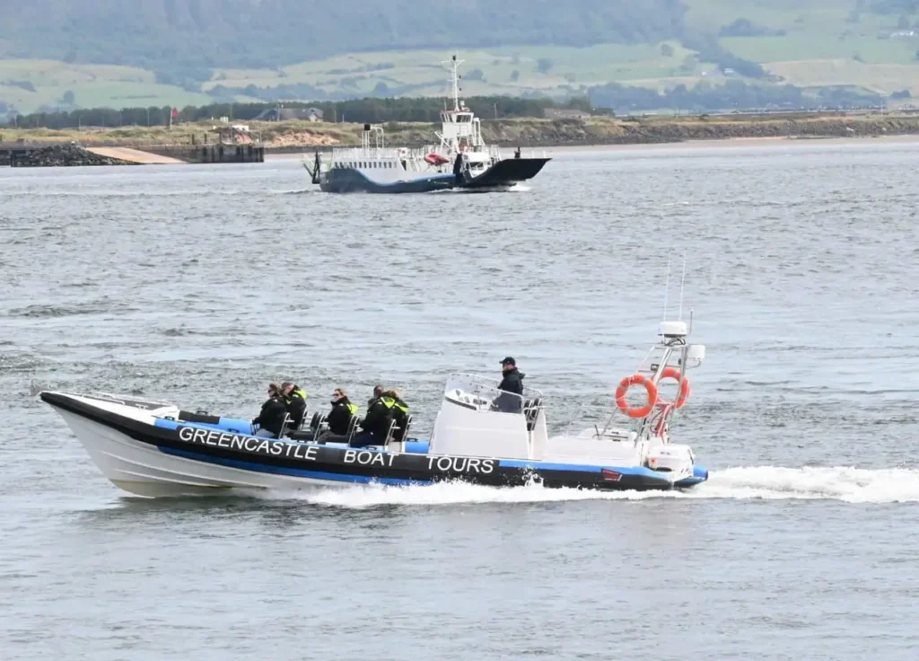 Greencastle Boat Tours RIB speeds across Lough Foyle; passengers in lifejackets, ferry and green hills behind.