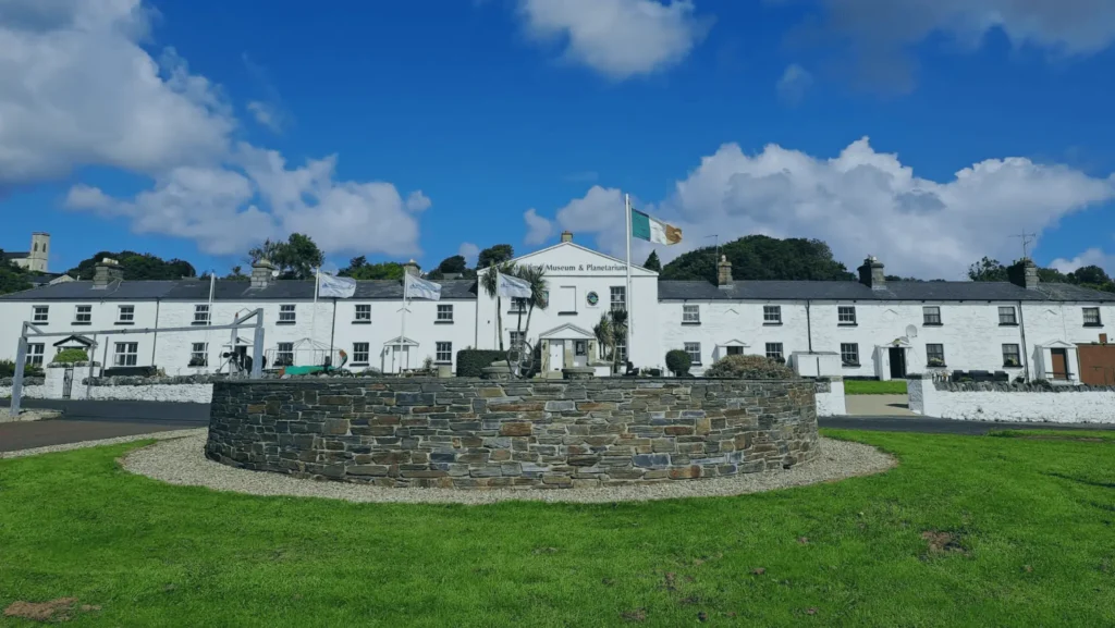 Irish flag flying over a white museum and planetarium, with a stone circular wall and green lawn under a bright blue sky