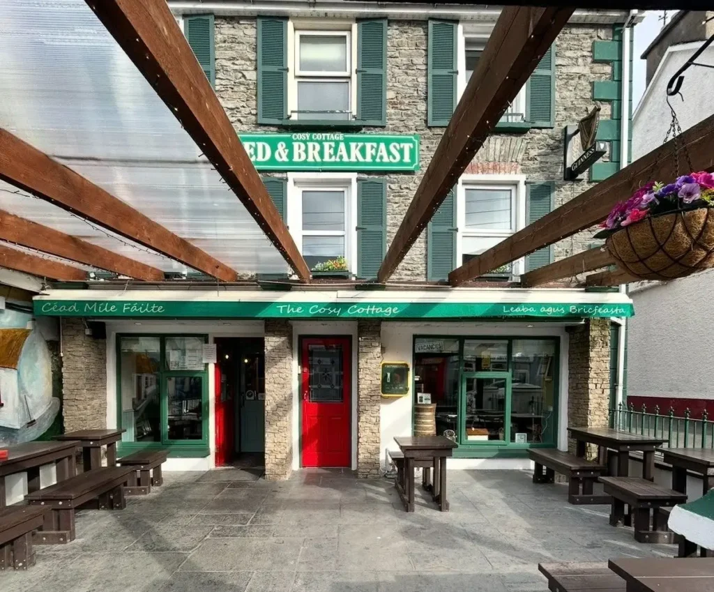 The Cosy Cottage B&B courtyard with outdoor benches, stone front, green shutters and a red door under wooden beams.