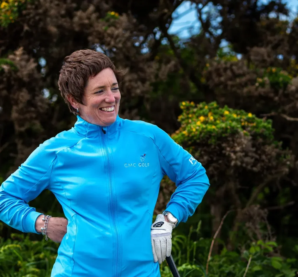 Smiling golfer in a blue jacket holding a club, standing outdoors with lush greenery in the background.