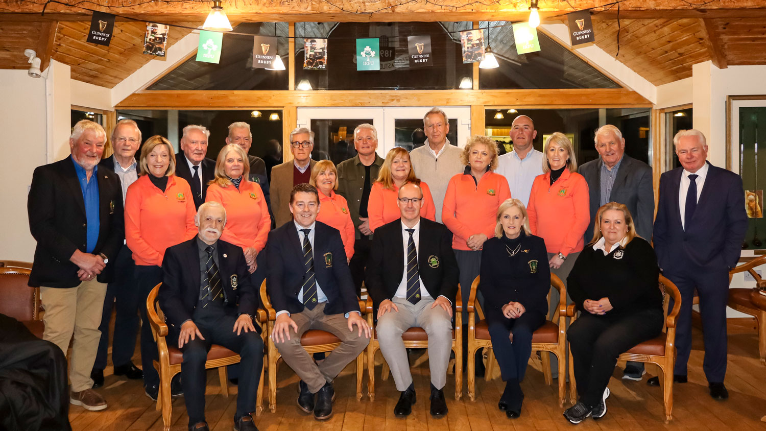 A group of people, some in orange jackets, are gathered indoors, sitting and standing, under rugby banners.